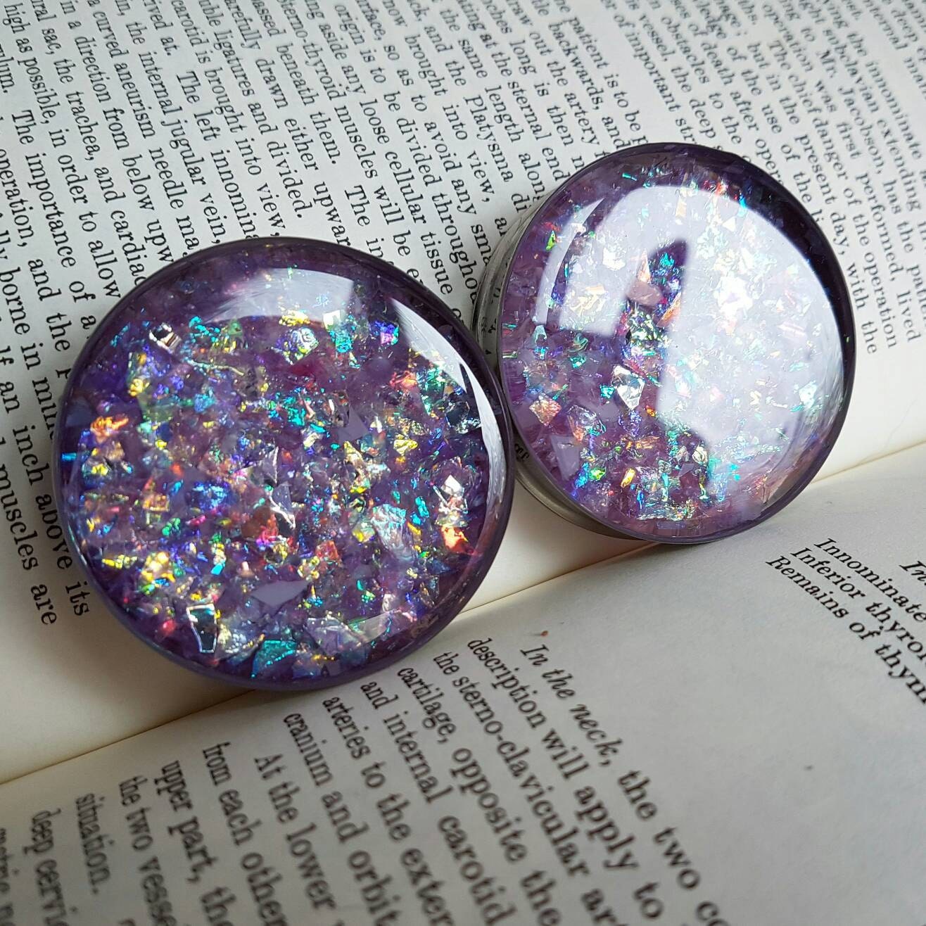 The image shows some large plugs for stretched ears onthe background of book. The plugs themselves are made from bright purple glitter in a stainless steel plug.