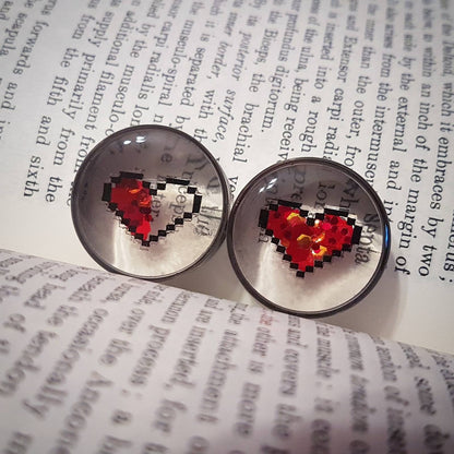 The image shows our 8 bit heart plugs sitting against a book backdrop, the plugs themselves are mostly transparent but you can see the 8 bit heart design set in the middle. The hearts are hand drawn in a 8bit style and red glitter is usedto fill them