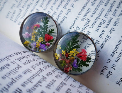 The photo shows our pressed flower plugs on a book backdrop, the plugs showcase The design where ferns and pressed flowers have been put in the plug in a V shape starting at the bottom with a transparent background. They come with multiple colours.