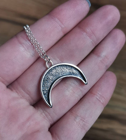 Silver crescent moon necklace held in a hand against a blurred background