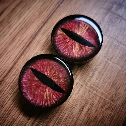 Photo shows a large pair of our dragon eye plugs on a wood backdrop. The plugs are a mix of orange red and gold with a painted pupil in the middle to give the effect of a dragon eye in a stainless steel plug.