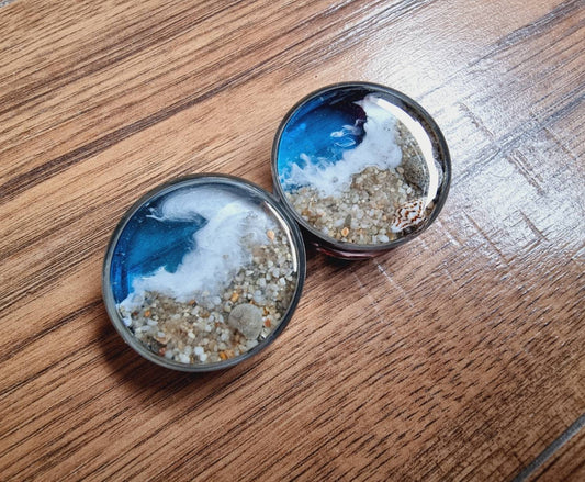 Plug on a wood backdrop, each stainless steel plug has the design built into it, for this we have clear blue resin on one side and sand on the other. Down the middle we have white hand painted waves that give the idea of an ocean in motion