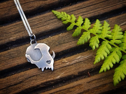 Silver cat skull pendant necklace on a wooden surface with green leaves