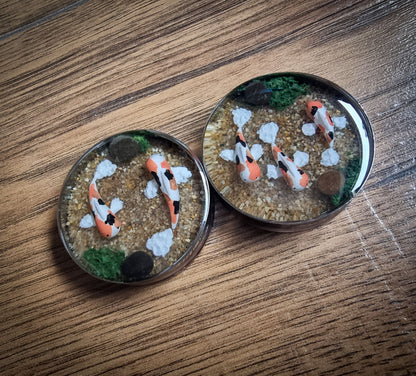 Two plugs for stretched ears with koi fish and gravel on a wooden surface