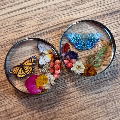 Two round plug earrings with butterflies and flowers on a wooden surface