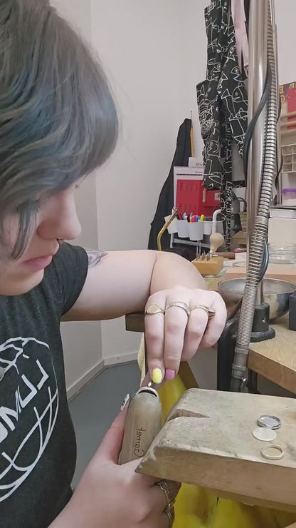 A short video showing the maker making a ring, its a small shot of a purple haired woman hand engraving a silver ring in a ring clamp. this goes on to show the finished ring, a wide band with hand engraved stars, rotating to show the frosted finish and mixed stars 